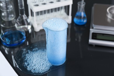 Photo of Beaker with blue sample, other laboratory glassware and scale on black mirror table, closeup