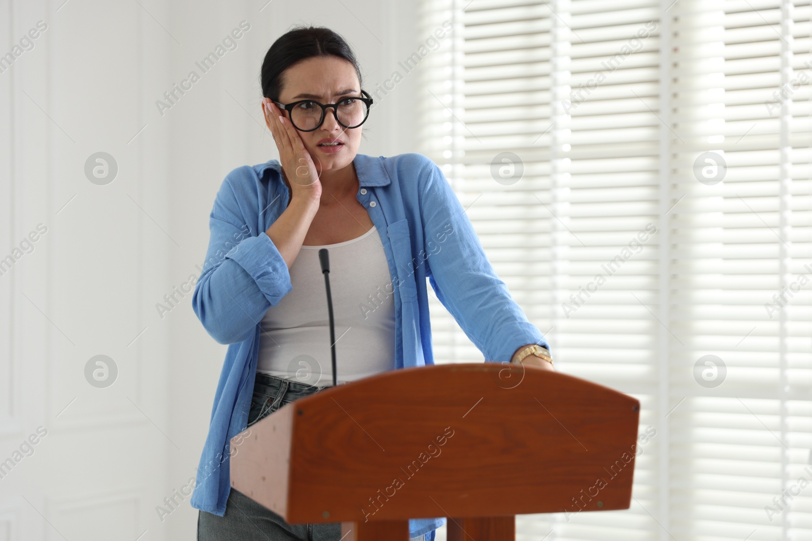 Fear of public speaking. Nervous woman with microphone at lectern indoors Photo of Fear of public speaking. Nervous woman with microphone at lectern indoors