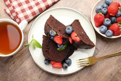 Photo of Pieces of delicious chocolate bundt cake with strawberries, blueberries and mint served on wooden table, flat lay