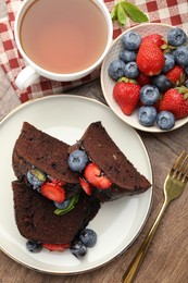 Photo of Pieces of delicious chocolate bundt cake with strawberries and blueberries served on wooden table, flat lay