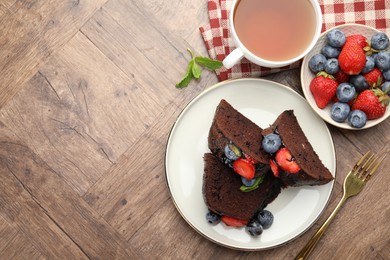 Photo of Pieces of delicious chocolate bundt cake with strawberries and blueberries served on wooden table, flat lay. Space for text