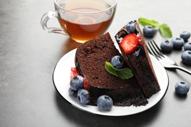 Photo of Pieces of delicious chocolate bundt cake with strawberries, mint and blueberries on grey table, closeup