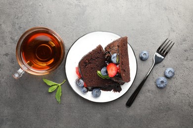 Photo of Pieces of delicious chocolate bundt cake with strawberries, mint and blueberries served on grey textured table, flat lay
