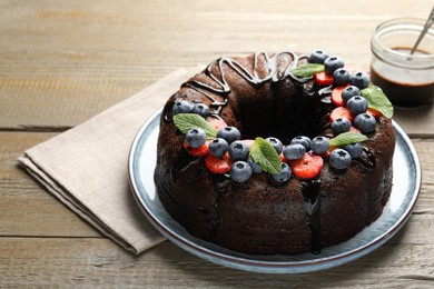 Photo of Delicious chocolate bundt cake with strawberries, mint and blueberries on wooden table, closeup