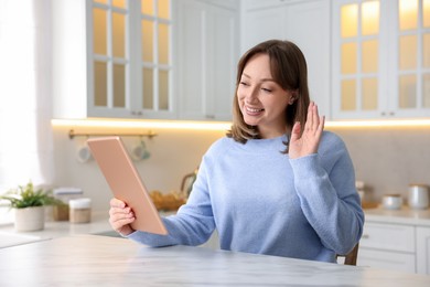 Photo of Woman having video chat via modern tablet at white marble table in kitchen