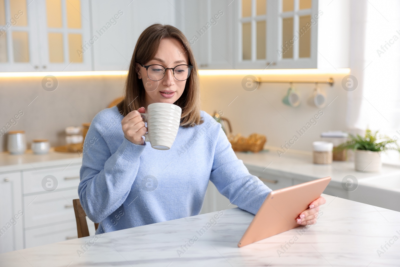 Woman with modern tablet and cup of drink at white marble table in kitchen Photo of Woman with modern tablet and cup of drink at white marble table in kitchen