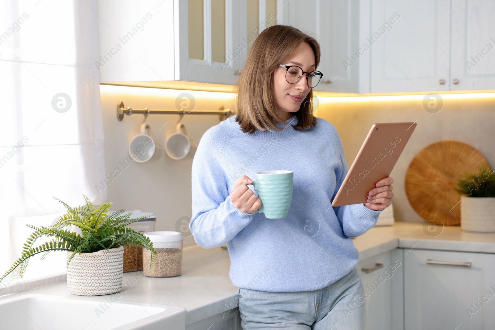 Woman with modern tablet and cup of drink in kitchen Photo of Woman with modern tablet and cup of drink in kitchen