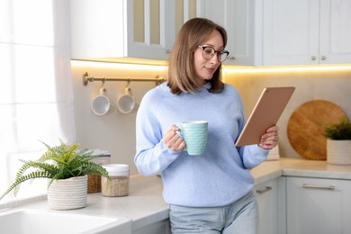 Photo of Woman with modern tablet and cup of drink in kitchen