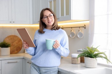 Photo of Woman with modern tablet and cup of drink in kitchen
