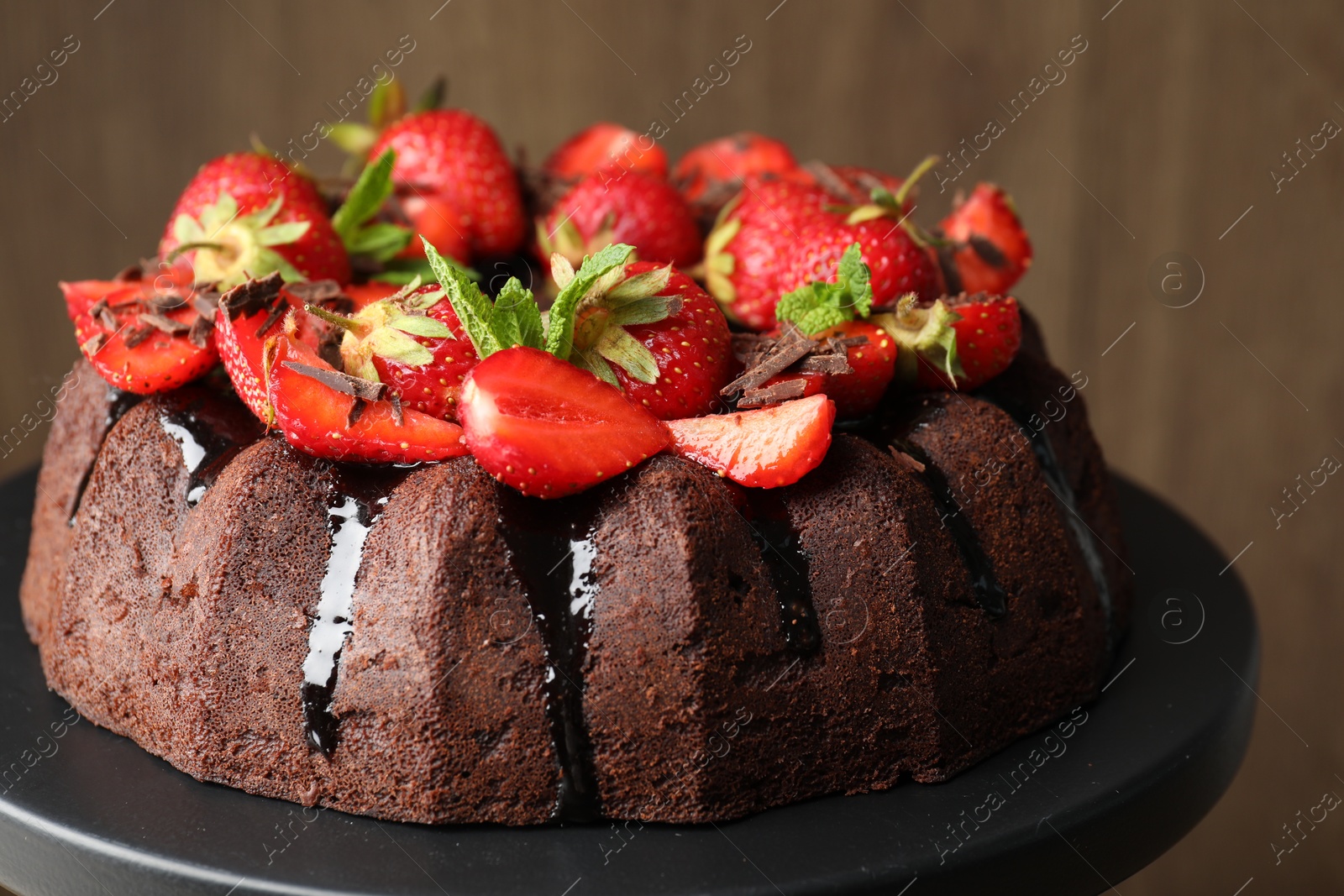 Delicious bundt cake with strawberries, mint and chocolate shavings on stand against blurred background, closeup Photo of Delicious bundt cake with strawberries, mint and chocolate shavings on stand against blurred background, closeup