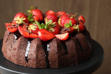 Photo of Delicious bundt cake with strawberries, mint and chocolate shavings on stand against blurred background, closeup