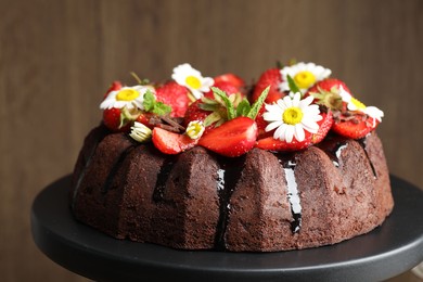 Photo of Delicious chocolate bundt cake with strawberries, chamomile flowers and mint on stand against blurred brown background, closeup