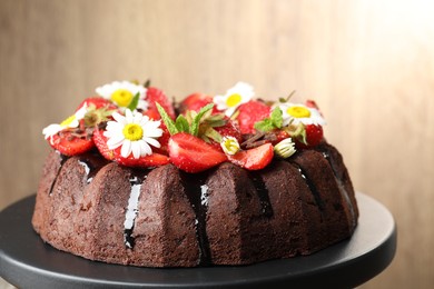 Photo of Delicious chocolate bundt cake with strawberries, chamomile flowers and mint on stand against blurred beige background, closeup