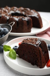 Photo of Piece of delicious chocolate bundt cake with mint and blueberries on light grey textured table against black background, closeup