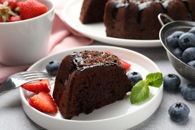 Photo of Piece of delicious chocolate bundt cake with strawberries, mint and blueberries on light grey textured table, closeup