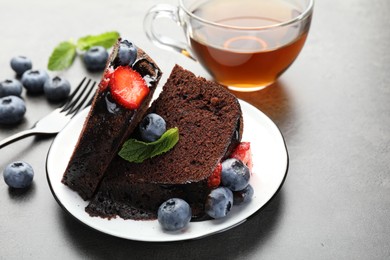 Photo of Pieces of delicious chocolate bundt cake with strawberries, mint and blueberries on grey table, closeup