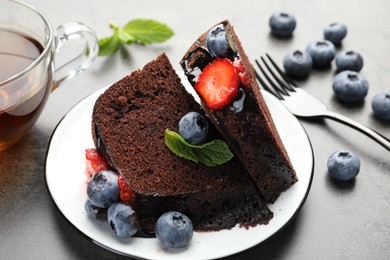 Photo of Pieces of delicious chocolate bundt cake with strawberries, mint and blueberries on grey table, closeup