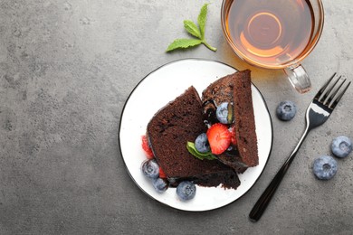 Photo of Pieces of delicious chocolate bundt cake with strawberries, mint and blueberries served on grey table, flat lay. Space for text