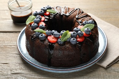 Photo of Delicious chocolate bundt cake with strawberries, mint and blueberries on wooden table, closeup