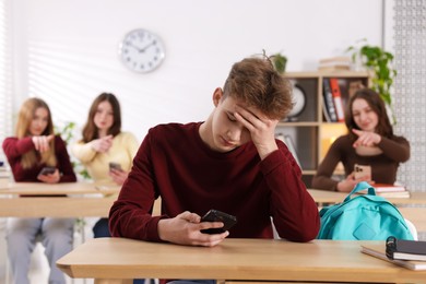Photo of Cyber bullying. Upset teenage boy with smartphone and schoolmates pointing at him in classroom, selective focus