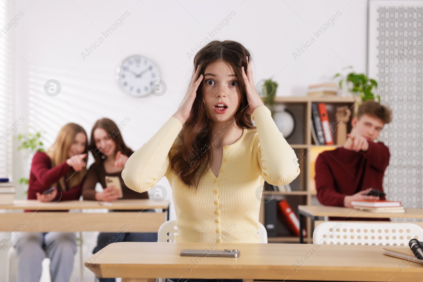 Cyber bullying. Upset teenage girl with smartphone and schoolmates pointing at her in classroom, selective focus Photo of Cyber bullying. Upset teenage girl with smartphone and schoolmates pointing at her in classroom, selective focus
