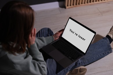 Photo of Cyber bullying. Upset teenage girl looking at laptop with text You Loser on floor indoors, closeup