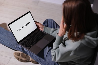 Photo of Cyber bullying. Upset teenage girl looking at laptop with text You Loser on floor indoors, closeup