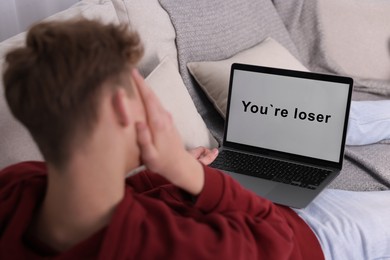 Photo of Cyber bullying. Upset teenage boy looking at laptop with text You Loser on sofa at home, closeup