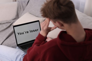 Photo of Cyber bullying. Upset teenage boy looking at laptop with text You Loser on sofa at home, closeup
