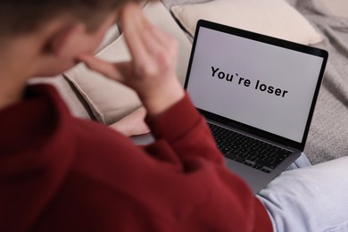 Photo of Cyber bullying. Upset teenage boy looking at laptop with text You Loser on sofa at home, closeup
