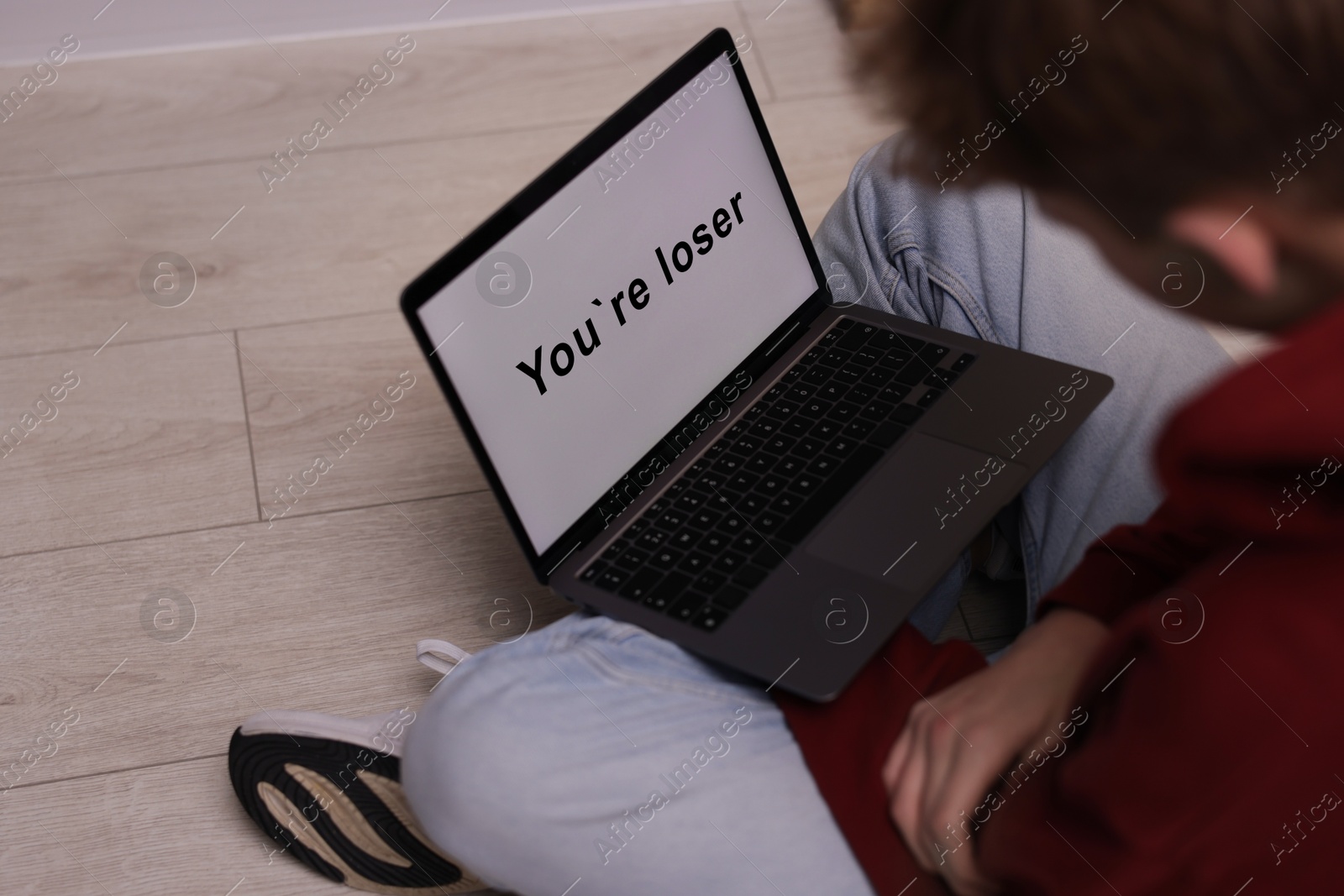 Cyber bullying. Upset teenage boy looking at laptop with text You Loser on floor indoors, closeup Photo of Cyber bullying. Upset teenage boy looking at laptop with text You Loser on floor indoors, closeup