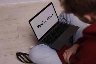 Photo of Cyber bullying. Upset teenage boy looking at laptop with text You Loser on floor indoors, closeup