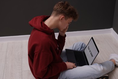 Photo of Cyber bullying. Upset teenage boy looking at laptop with text You Loser on floor indoors