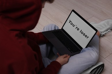 Photo of Cyber bullying. Upset teenage boy looking at laptop with text You Loser on floor indoors, closeup