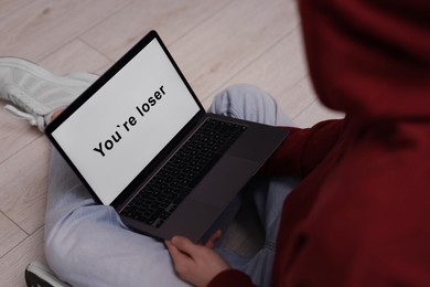 Photo of Cyber bullying. Upset teenage boy looking at laptop with text You Loser on floor indoors, closeup