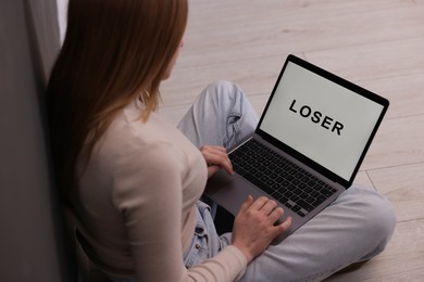 Photo of Cyber bullying. Upset teenage girl looking at laptop with text You Loser on floor indoors, closeup