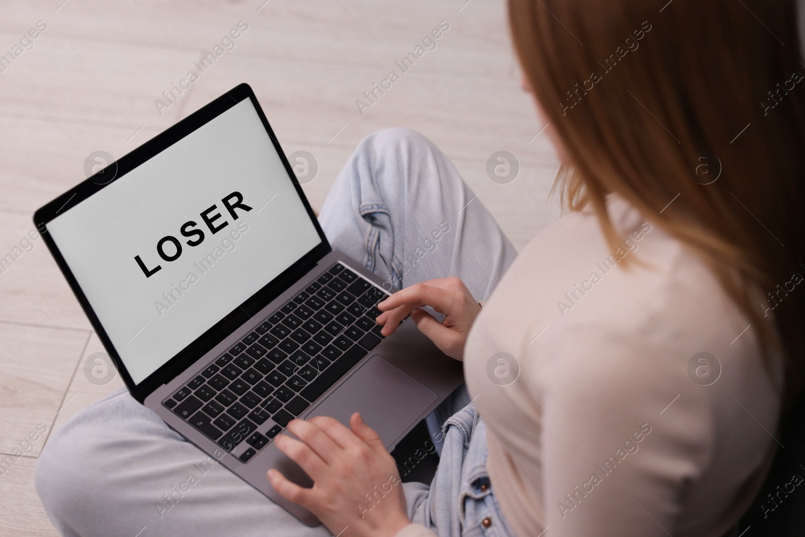 Cyber bullying. Upset teenage girl looking at laptop with text You Loser on floor indoors, closeup Photo of Cyber bullying. Upset teenage girl looking at laptop with text You Loser on floor indoors, closeup
