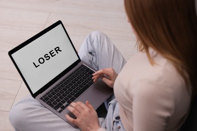 Photo of Cyber bullying. Upset teenage girl looking at laptop with text You Loser on floor indoors, closeup