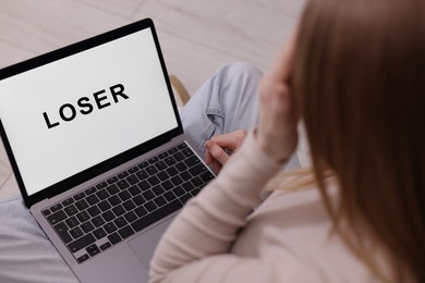 Photo of Cyber bullying. Upset teenage girl looking at laptop with text You Loser on floor indoors, closeup