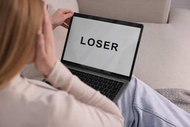 Photo of Cyber bullying. Upset teenage girl looking at laptop with text You Loser on sofa at home, closeup