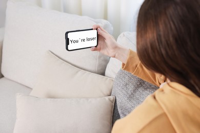 Photo of Cyber bullying. Upset teenage girl looking at smartphone with text You Loser on sofa at home, closeup