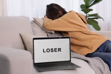 Photo of Cyber bullying. Upset teenage girl and laptop with text You Loser on sofa at home, selective focus