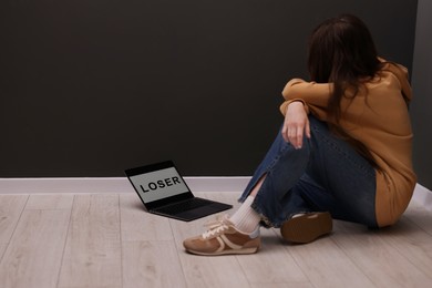 Photo of Cyber bullying. Upset teenage girl and laptop with text You Loser on floor indoors. Space for text
