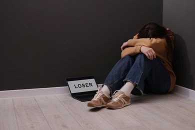 Photo of Cyber bullying. Upset teenage girl and laptop with text You Loser on floor indoors. Space for text