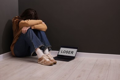 Photo of Cyber bullying. Upset teenage girl and laptop with text You Loser on floor indoors. Space for text