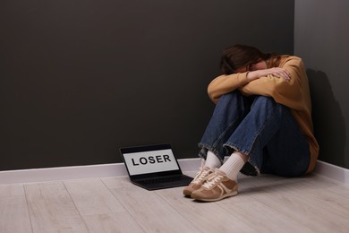 Photo of Cyber bullying. Upset teenage girl and laptop with text You Loser on floor indoors. Space for text