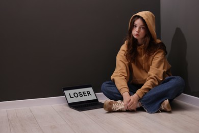 Photo of Cyber bullying. Upset teenage girl and laptop with text You Loser on floor indoors. Space for text