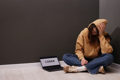 Photo of Cyber bullying. Upset teenage girl and laptop with text You Loser on floor indoors. Space for text