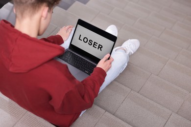 Photo of Cyber bullying. Upset teenage boy looking at laptop with text You Loser on stairs indoors, closeup