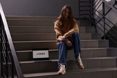 Photo of Cyber bullying. Upset teenage girl and laptop with text You Loser on stairs indoors